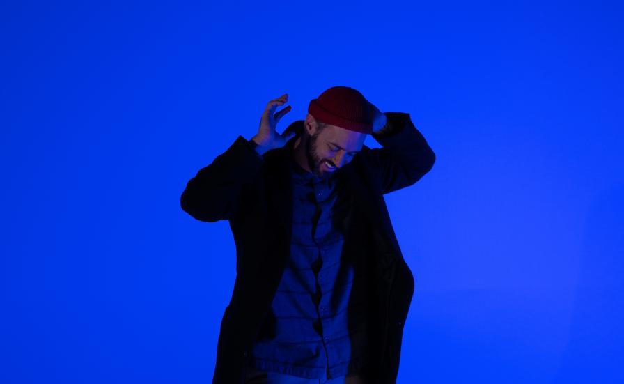 Ryan Mason standing, he is smiling and raising his hands abowe his head, looking to the floor. Wearing a red cap and a black coat. The backround is bright dark blue.