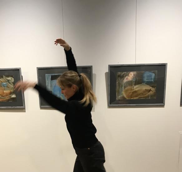Kaisa Hahl dancing in front of pictures on the wall. Her hands are lifted up.