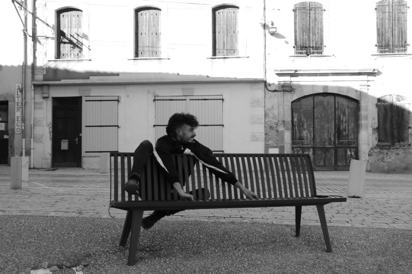 Raphaël Beau in a black and white picture sitting on a park pench with a white building on the backround.